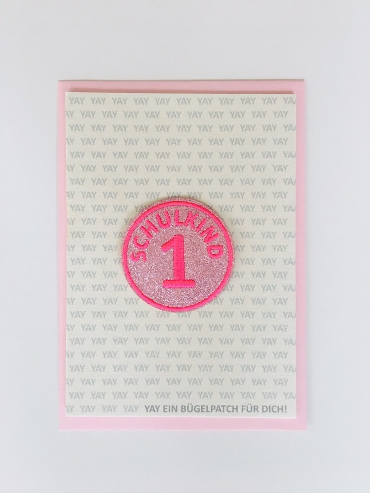 0D88A7E4-813B-4C96-A119-B70D88A604A6 Bügelpatch, rund, rosa, Glitzer, Aufschrift „Schulkind 1“, Hintergrund Schriftzug „Yay“, Papierkarte, rosa Rand.