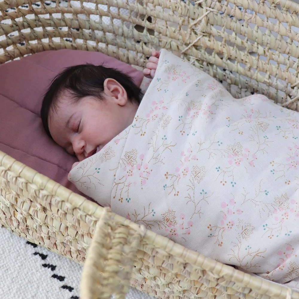 Babykorb aus geflochtenem Material, lila Kissen, Baby in weißer Decke mit floralem Muster.