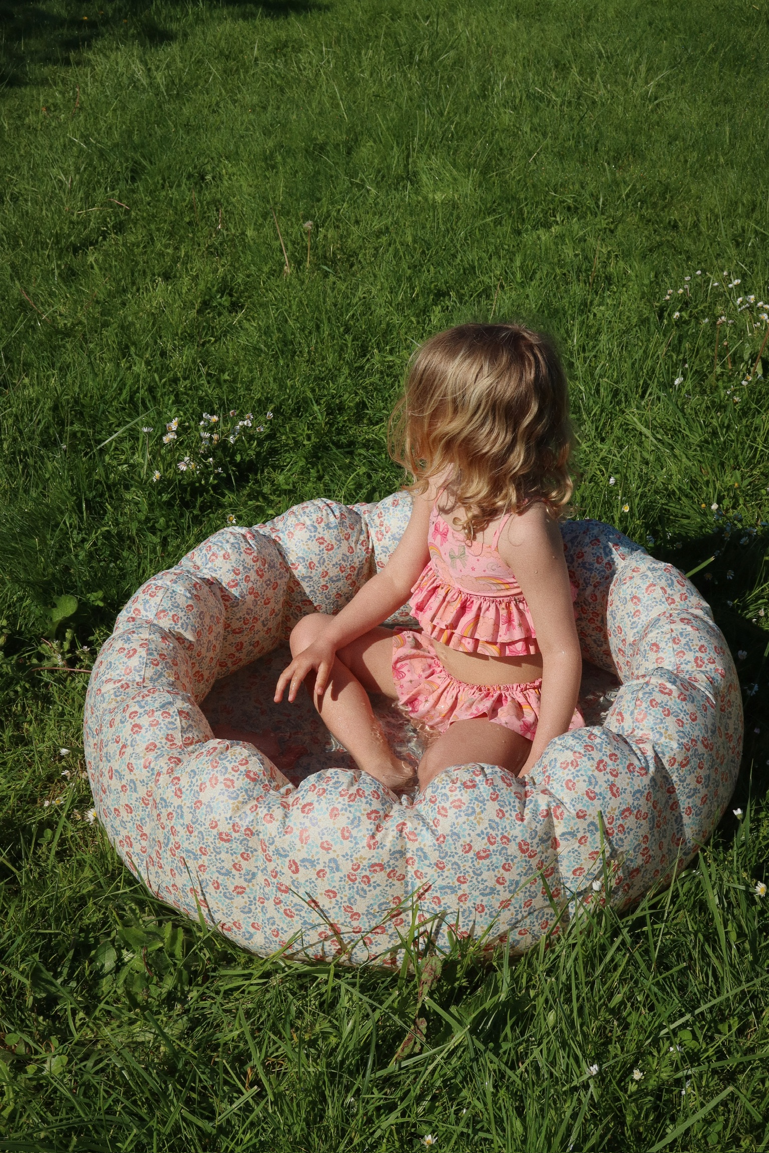 Aufblasbares Kinderspielbecken, blumiges Muster, rosa Badekleidung, sonniger Rasen.