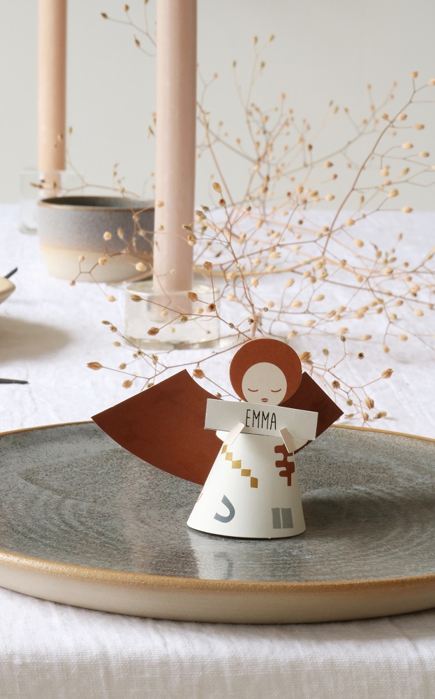 fridur-new-31 Engelfigur aus Papier, weiß-braun, rotes Haar, hält Schild mit "Emma", beige-grauer Teller im Hintergrund.