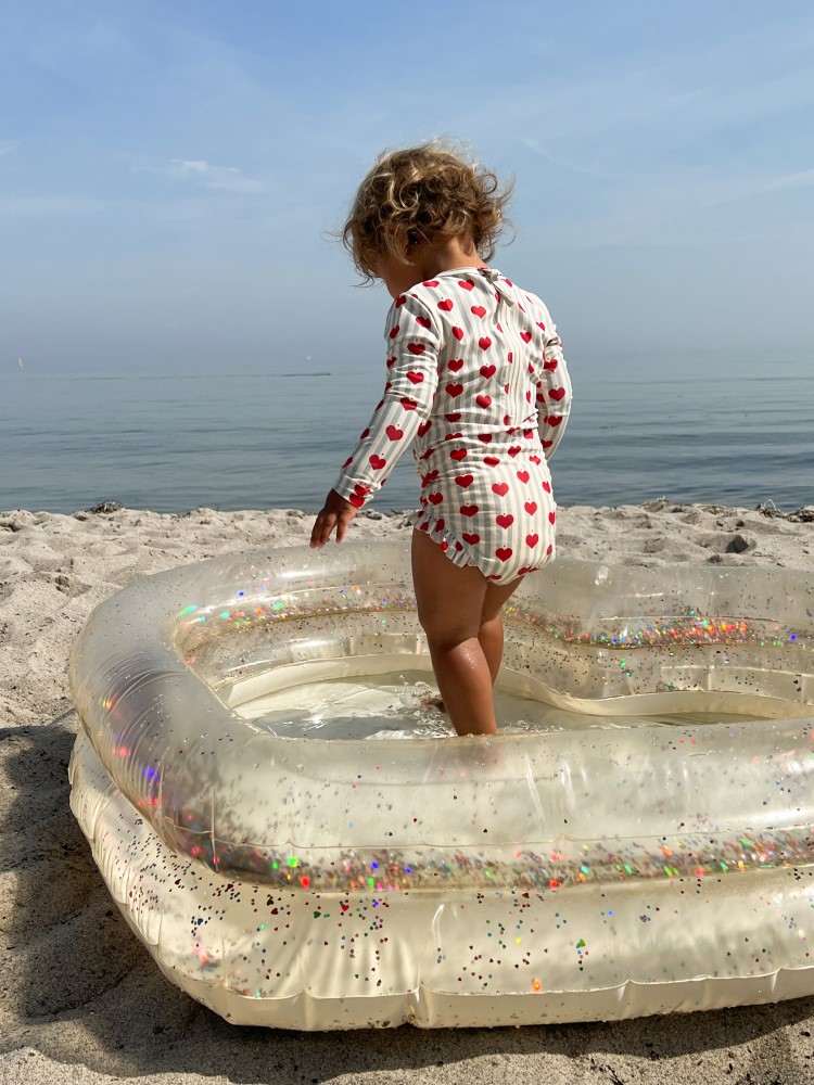 KS5195-HEART-POOL-TRANSPARENT-CREAM-Extra-3 Aufblasbarer Kinderpool, transparent mit buntem Glitzer; Kind in weißem Badeanzug mit roten Herzmustern.