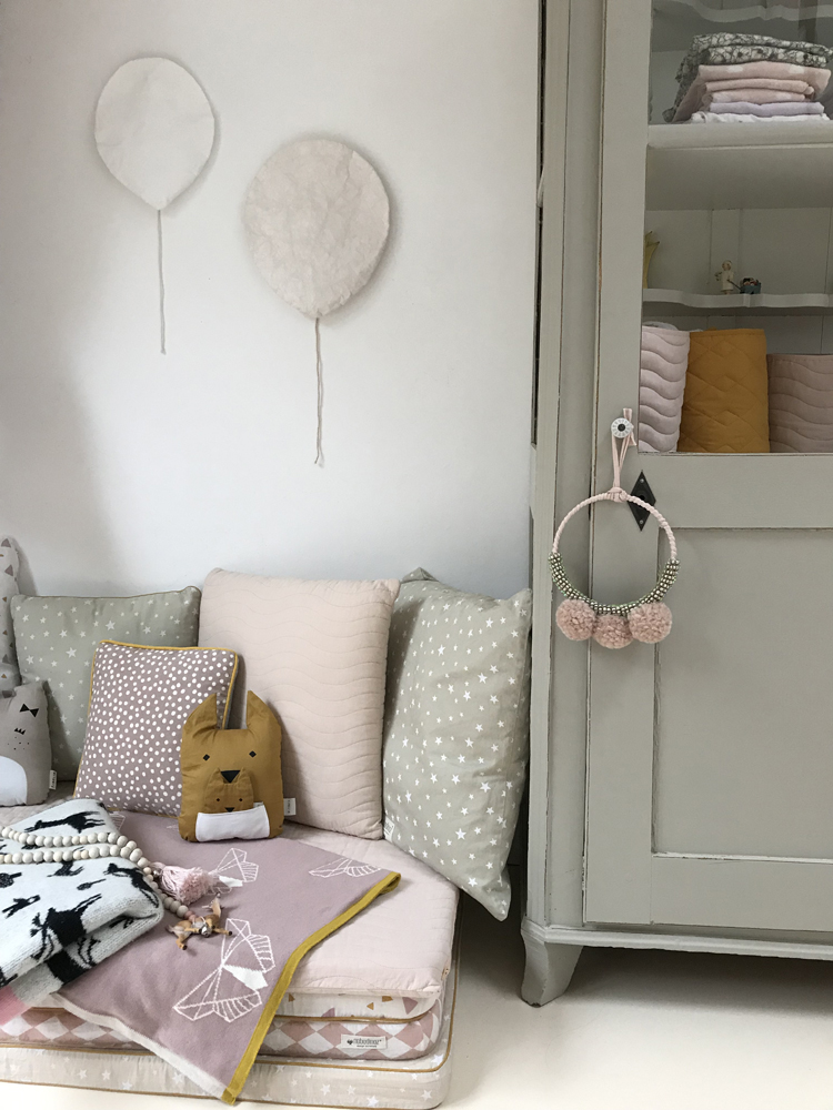 IMG_5823 Kissen in Beige mit Wellenmuster, Polster in Grün mit weißen Sternen, dekorative Kissen mit Tiergesicht, zwei Ballon-Wanddekorationen in Weiß, grauer Schrank mit gefalteter Bettwäsche, Holzperlenkette, pastellfarbene Decke mit grafischem Muster.