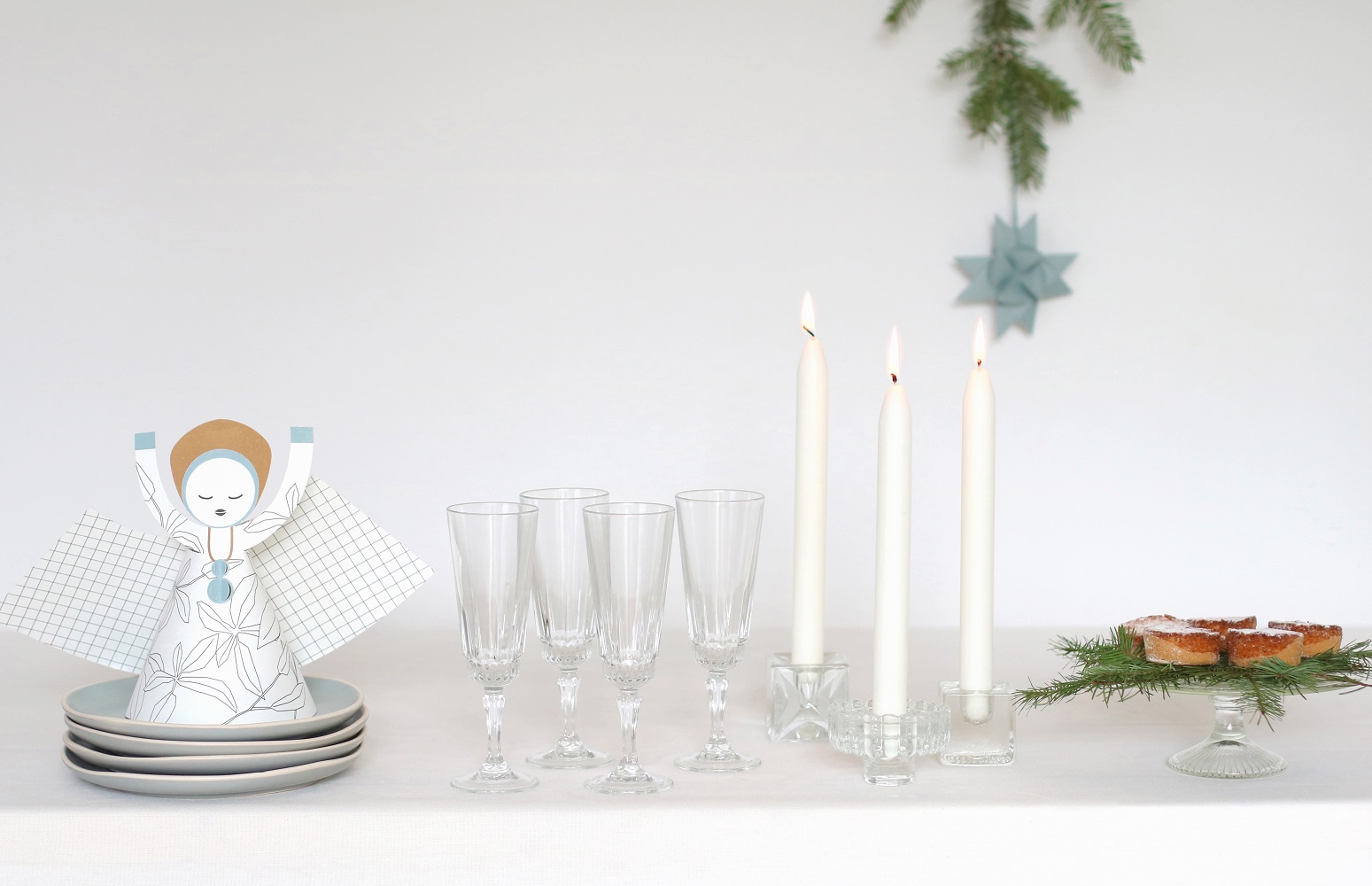 XL-angel-tablesetting-3 Kerzenhalter aus Glas, weiße Stabkerzen, Kristallgläser, Papierserviettenhalter in Engelsform, blauer und weißer Aufdruck, Kuchenständer mit Gebäck, Tannenzweig-Dekoration.