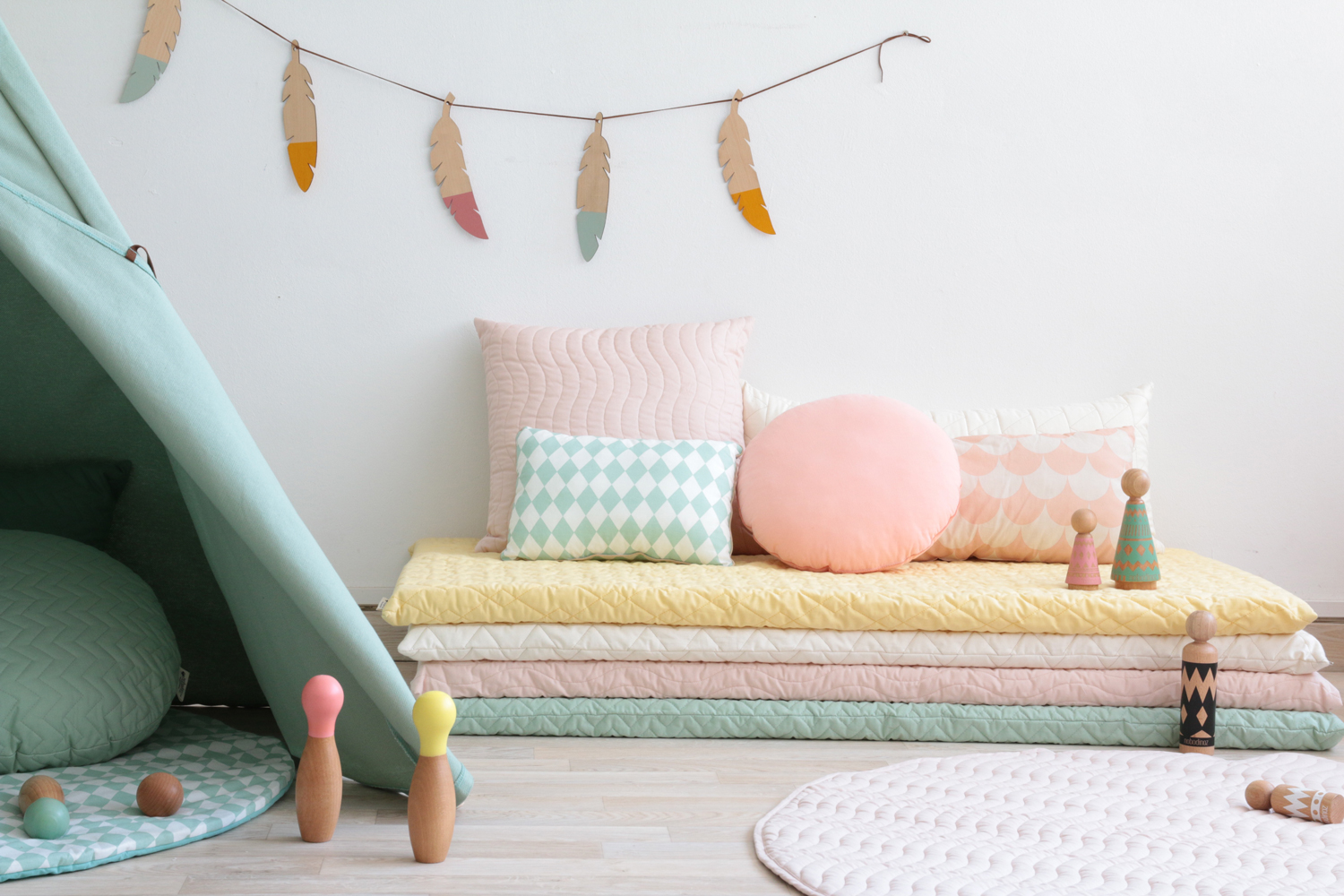 teepee-nevada-provence-green-monaco-matress-cushions-quilted-nobodinoz Kinderzimmerdeko, Spielzeugkegel in Pastellfarben, übereinander gestapelte Matratzen in Gelb, Creme, Rosa, Mint; Kissen mit Rautenmuster, Feder-Girlande; Zelt in Mint.