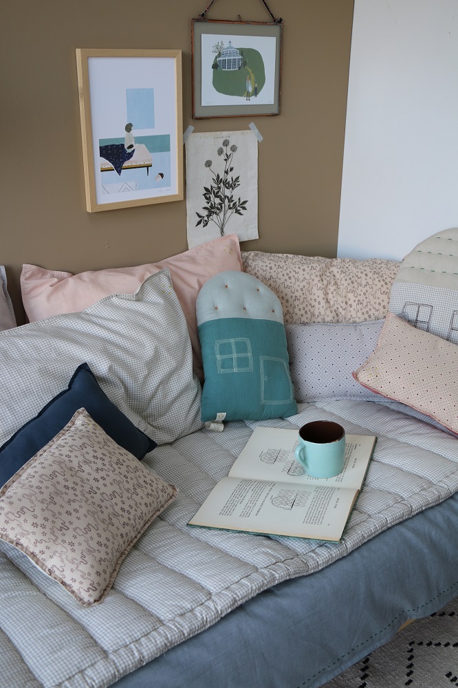 detail28 Sofa mit gemusterten Kissen in Beige, Blau und Mint; Patchwork-Design; zwei gerahmte Wandbilder; Buch und mintfarbene Tasse auf Sofa; Kissen mit Häusern und abstrakten Mustern.