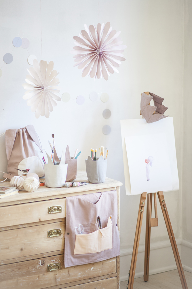 IMG_8517591b059bbe878 Holzkommode mit Schubladen; Organizerbeutel mit Taschen, rosa; Stoffbehälter gefüllt mit Stiften und Pinseln; Staffelei aus Holz, weißes Blatt mit gemaltem Motiv; Papierblumen in zartem Rosa und Weiß an der Wand.