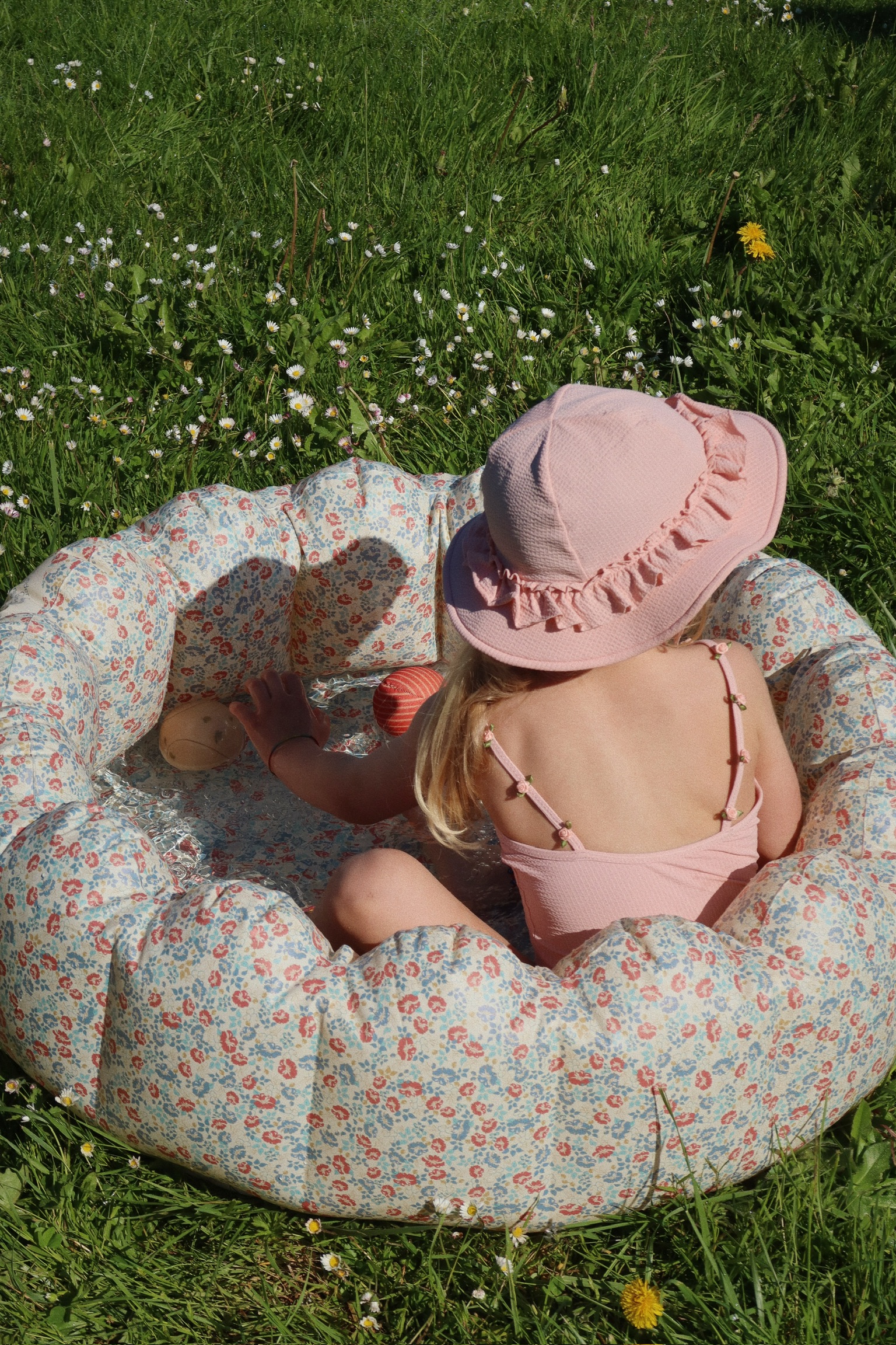 Aufblasbares Kinderplanschbecken, florales Muster, Mädchen in rosa Badeanzug und Sonnenhut, Spielzeugbälle im Wasser.
