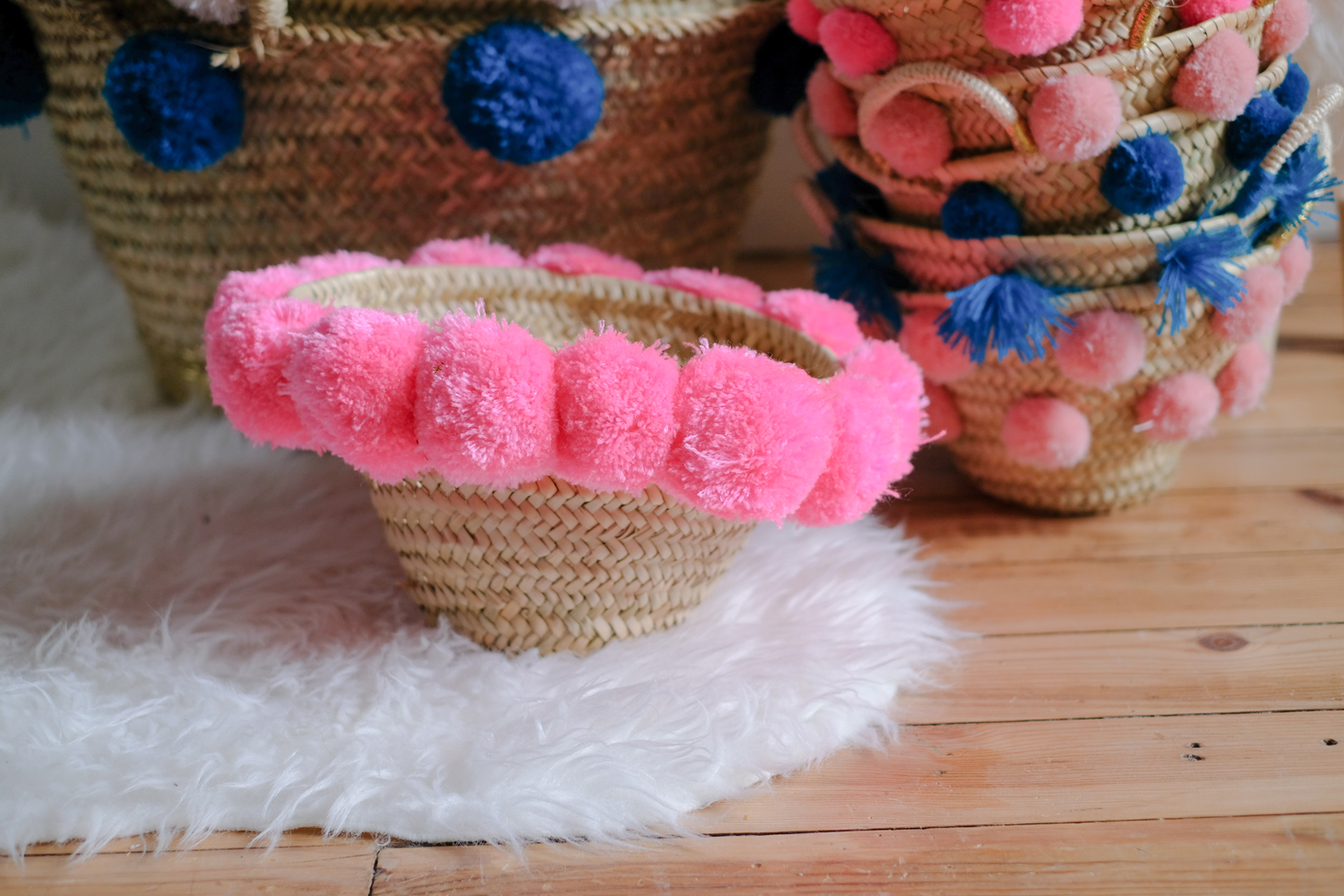 KYRLA_FLUOPINK_2 Korb aus geflochtenem Stroh, verziert mit pinken Pompoms; im Hintergrund weitere Körbe mit blauen und pinken Pompoms.