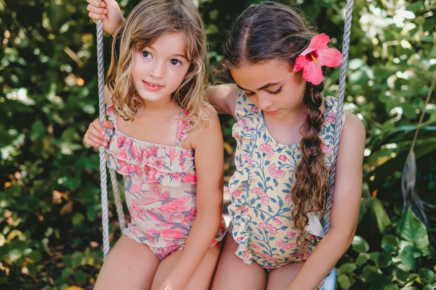 girls-bathing-suit-zacatecas-pink-flowers-2 Zwei Mädchen auf Schaukel, gemusterte Sommerkleider; links rosa Blütenmuster, Rüschen; rechts gelbes Kleid, florales Muster, rote Blume im Haar