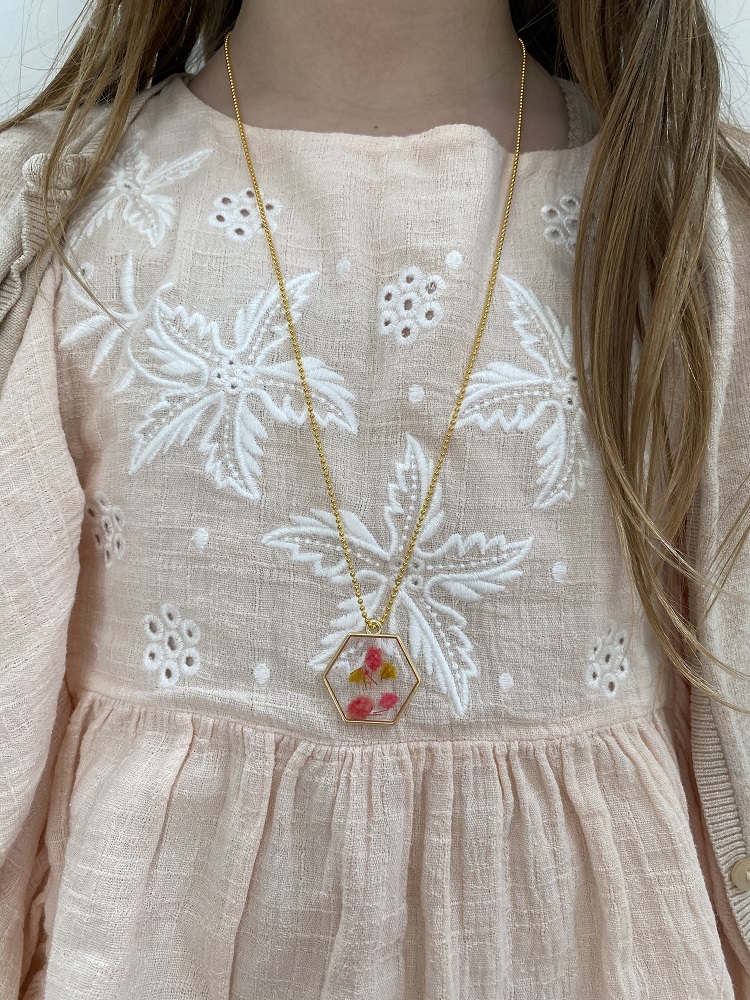 IMG_2610 Kette, goldene Farbe, sechseckiger Anhänger mit rosa und gelben Blumen, hellrosa Kleid mit weißem Stickmuster.