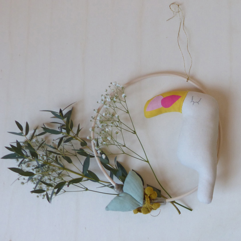 Scala-Charlie-baby-toucan-1 Wanddekoration, Stoff-Tukan, weiß, gelber Schnabel, rosa Akzent, runder Holzrahmen, dekorative Pflanzen, zarte Blüten.