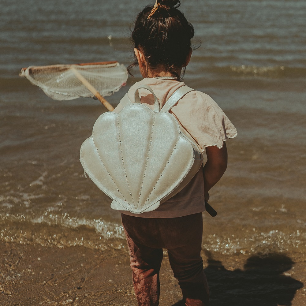 Bags-Purses-Crop-Media-kit-SS2212 Kinderrucksack, Muschelform, hellbraun, am Rücken eines Kindes, Strandhintergrund, Kescher in der Hand.