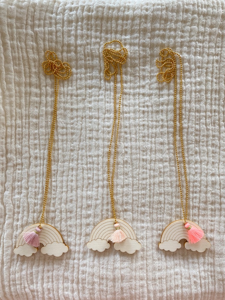 IMG_3091 Drei goldene Halsketten mit Holzanhängern in Regenbogenform, jeweils mit buntem Quastenanhang: rosa, creme und pink. Liegen auf weißem Stoffhintergrund.
