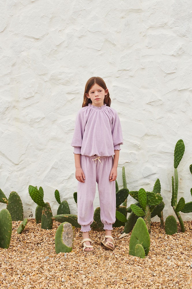 SS22_016_Terry-Smocked-Blouse_lavendervqvHBKRNdf4tW Kinderanzug, lila, weiches Material, langärmeliges Oberteil, elastische Bündchen, lange Hose, Kordelzug, Sandalen, neutraler Hintergrund.
