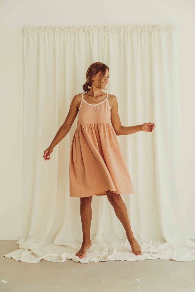 liss23_076_W_louisa-dress_peach_1-1 Kleid, ärmellos, hellorange, knieumspielend, ausgestellter Schnitt, weiße Träger, weibliches Model.