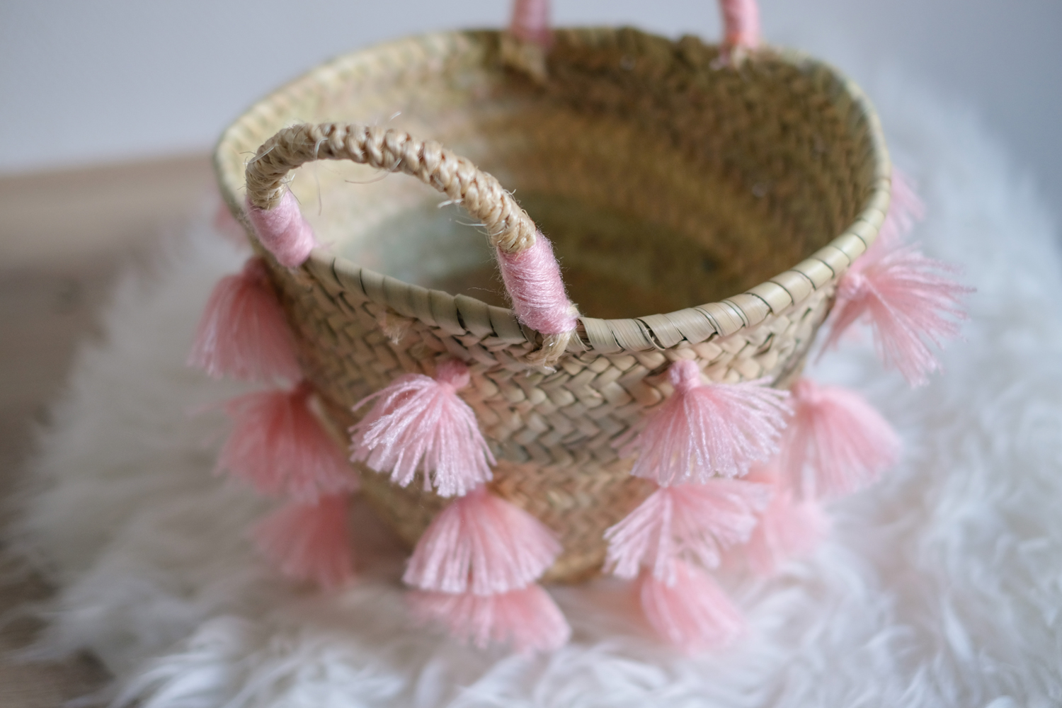 MOTI_BLOSSOM_3 Korb aus geflochtenem Naturmaterial, runde Form, Henkel mit rosa Garn umwickelt, rosa Quasten als Verzierung, cremefarbene Innenseite.