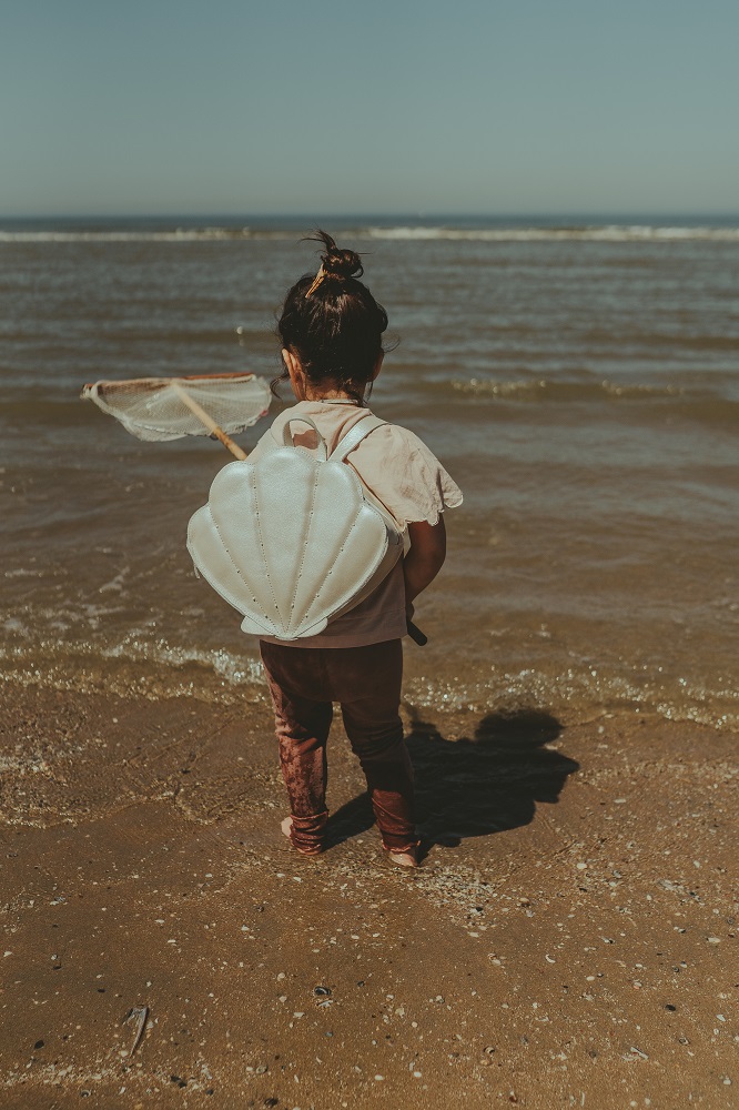 ANCK_donsjeHS2022123 Kind mit weißem Muschel-Rucksack am Strand, haltend ein Fischernetz, braune Kleidung.