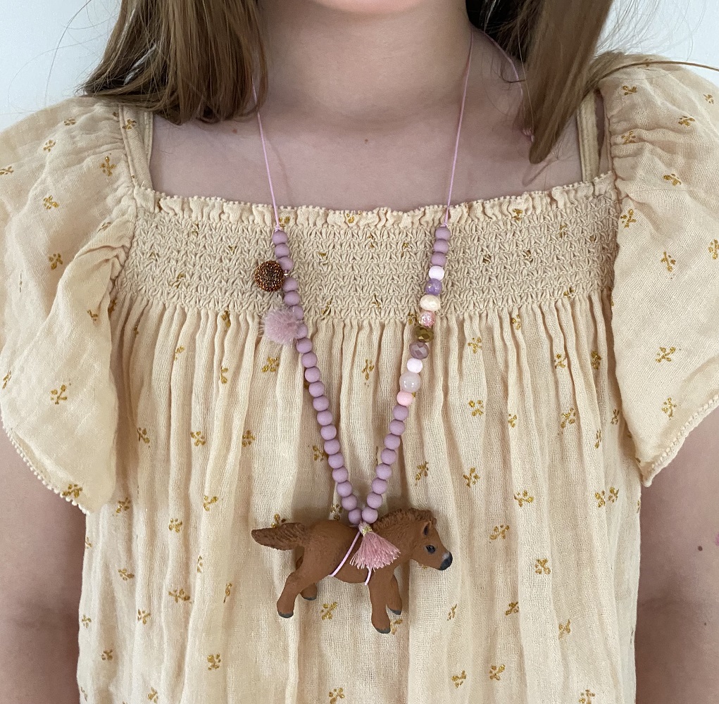 IMG_5072 Kette mit rosa Perlen, braunes Pferd-Anhänger, pastellfarbene Quaste, getragen auf cremefarbenem Kleid mit gelbem Blumenmuster.
