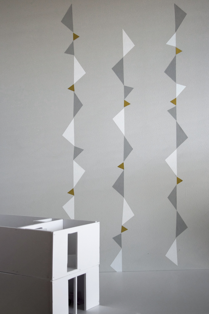 wallsticker-lines-01 Papierhausmodell weiß, geometrische Wanddeko; grau, weiß, goldene Dreiecke; vertikale Anordnung.
