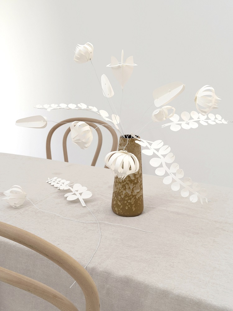 field-flowers-large-white-2 Vase mit braun-gesprenkelter Oberfläche, weiße filigrane Papierblumen und Blätter, auf beigefarbener Tischdecke, helle Holzstühle im Hintergrund.