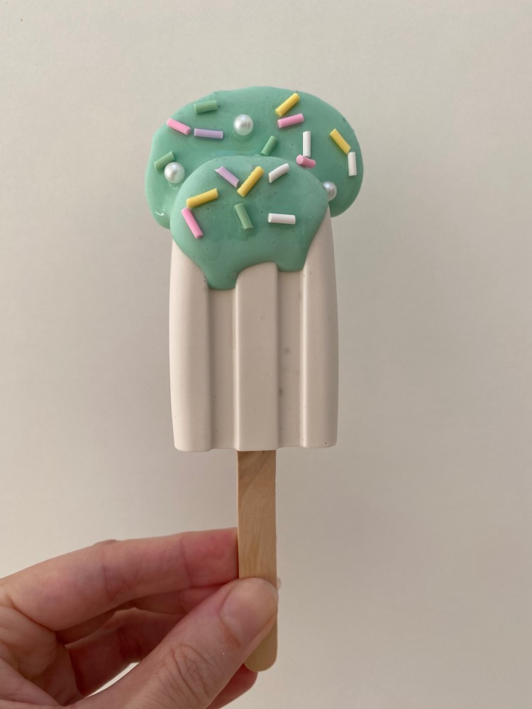 IMG_2494bekqn04llvReB Eisförmige Seife, Mintgrün-Weiße Glasur, bunte Zuckerstreusel, Holzstiel, Hand hält Produkt.