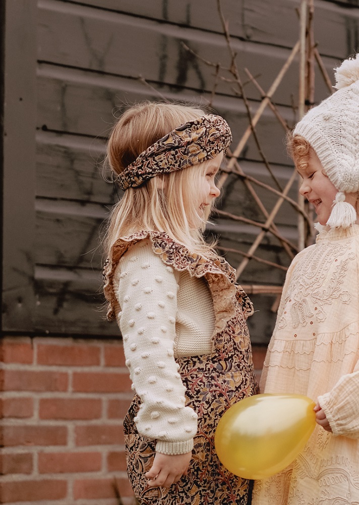 Girls-Headband-Daia-NordishFlowers-7 Kinderkleidung, Mädchen; links: Strickpullover beige mit Punkten, Latzhose gemustert, Haarband floral; rechts: cremefarbenes Kleid mit Spitze, Strickmütze weiß mit Bommel; gelber Luftballon.