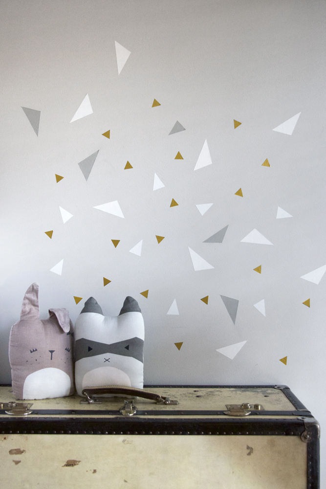 wallsticker-confetti Kissen in Tierform, links Hase, rosa, rechts Waschbär, grau-weiß; beide mit Gesichtsmotiven; Hintergrund weiße Wand mit goldenen, grauen und weißen Dreiecken; Kissen auf Vintage-Koffer platziert.