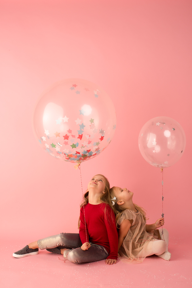AW2016-5 Zwei transparente Ballons mit bunten Sternen, zwei Kinder, eines in rotem Pullover und Jeans, das andere im beige Kleid, rosa Hintergrund.