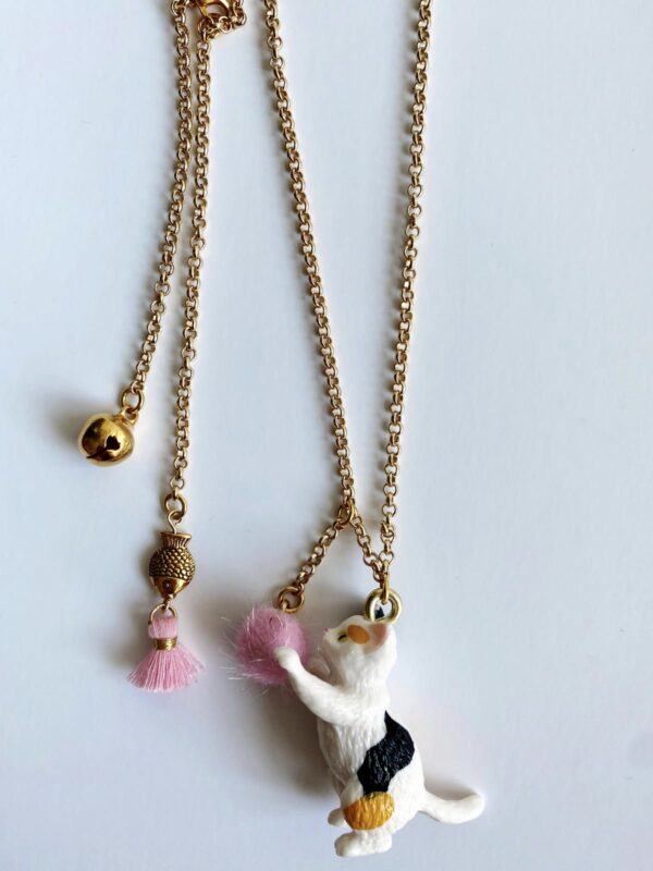 E63C65B6-9D33-43EA-A032-373C927CC1CB-600x800 Goldene Halskette, zwei Anhänger; erster: goldene Glocke; zweiter: weiße Katze mit schwarzem und orangefarbenem Fleck, pinker Pompon; zusätzlicher Anhänger: goldener Zylinder mit pinker Quaste.
