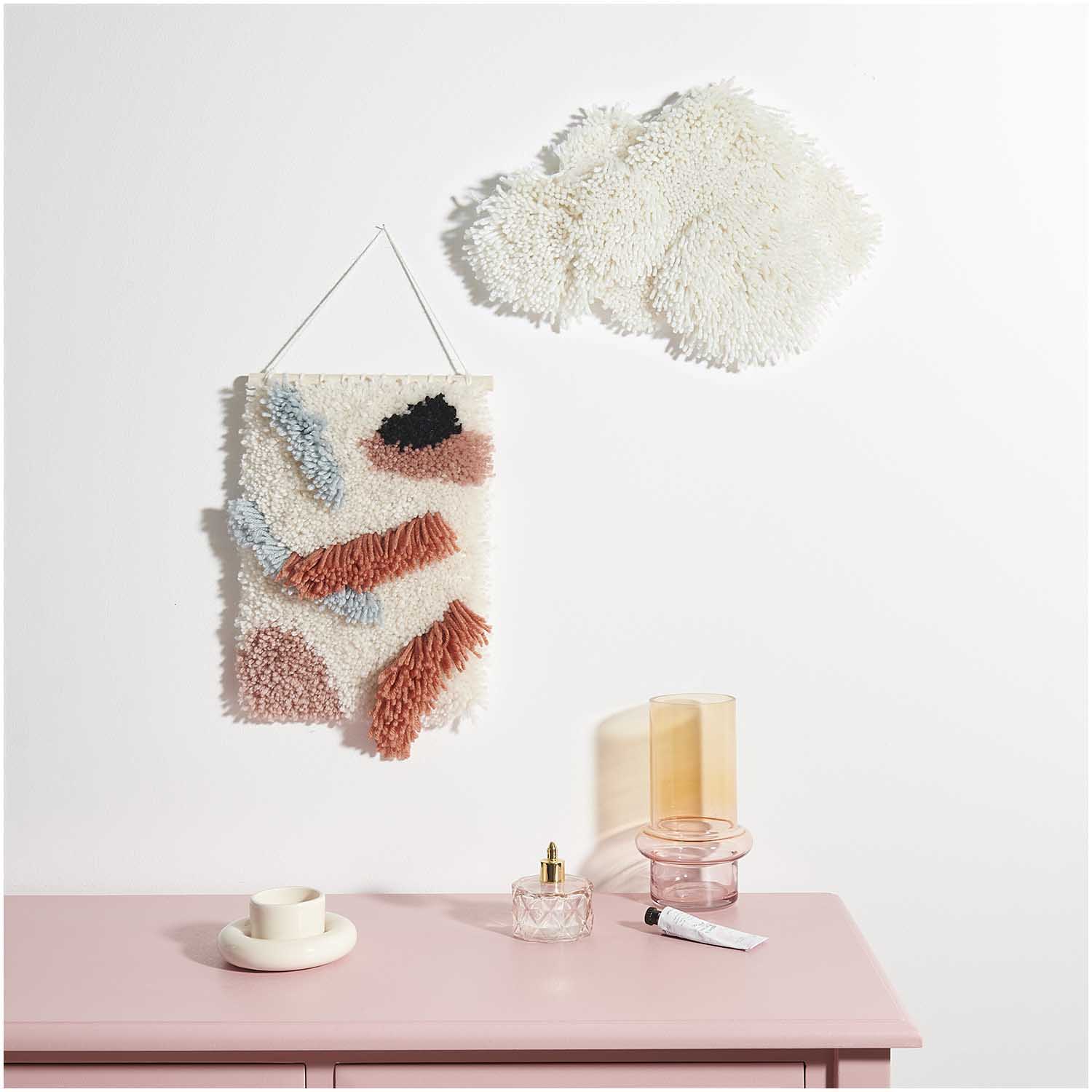 Wandteppich, bunte Fransen in Weiß, Blau, Rosa, Schwarz; daneben strukturierte weiße Wanddeko; rosafarbenes Möbel im Vordergrund mit Glasflakon, Teelicht, Tube, orange-rosa Glasvase.