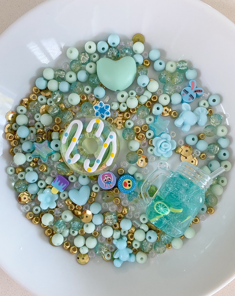 Perlen, türkis, gold, transparent; Herz, Donut, Glitzer; Knöpfe mit Tiermotiven; Miniaturglas mit blauer Flüssigkeit und Limettenscheibe; Runde und facettierte Varianten; Deko-Set auf weißem Teller.