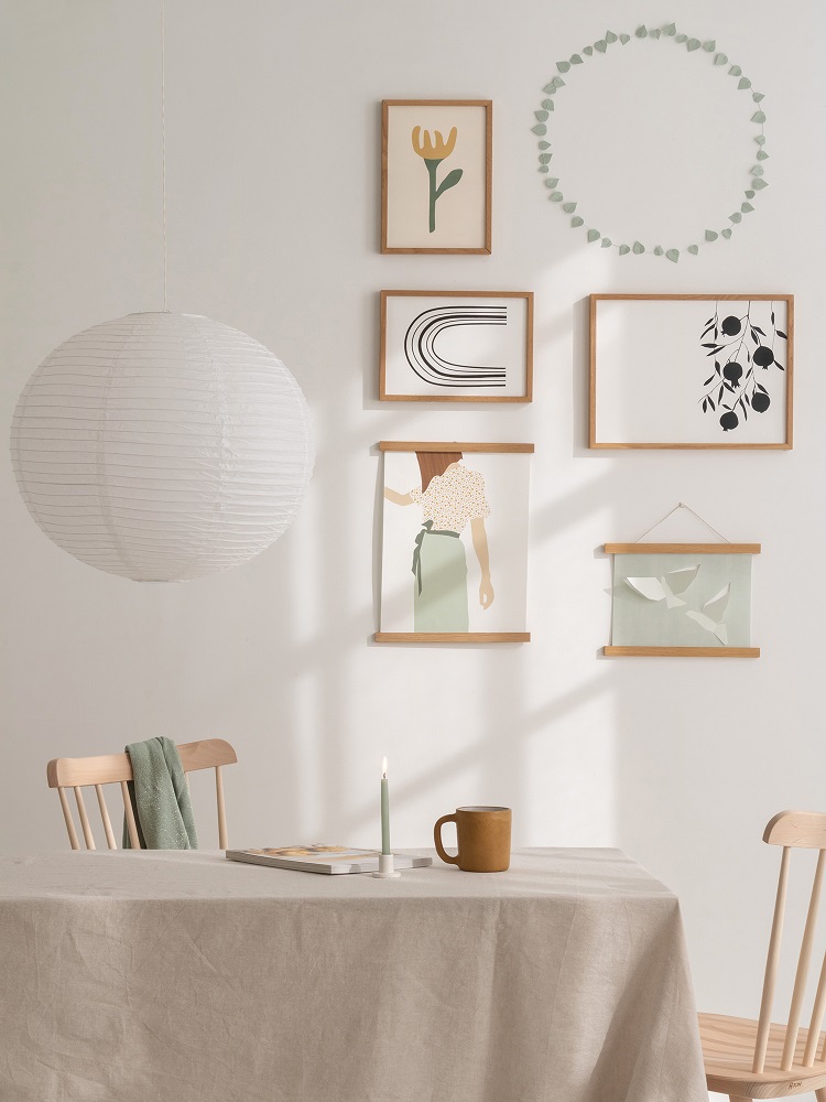 twig-leaves-sage-green-1 Holzrahmen mit Grafiken, runder Papierlampenschirm, beige Tischdecke, braune Keramiktasse, grünes Handtuch auf Stuhl, grüne Kerze im Halter auf Tisch, helle Wand.