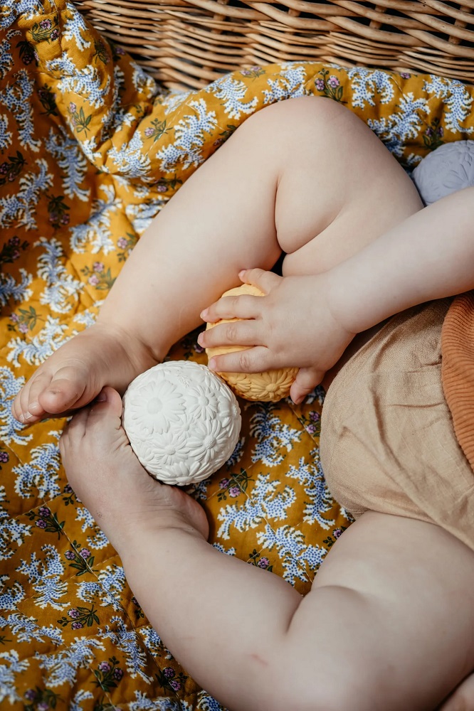 cn0rdem4tesq8fhhwhk0_f6a48beb-9076-4baa-8fdf-1626b0c4b48c_1024x1024-2x Baby, zwei Spielbälle, weiß und gelb, Blumenmuster-Aufdruck, gelbe Decke mit Blumenmotiv.