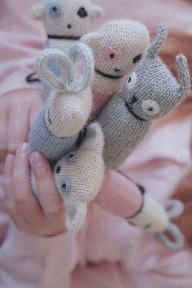 Minis Handpuppen, gestrickt, Tiermotive, Farben: beige, grau, rosa, schwarze Augen, schwarze Münder, Kind hält Puppen, detailreiche Gesichtszüge, weiche Textur, kindgerechtes Design.
