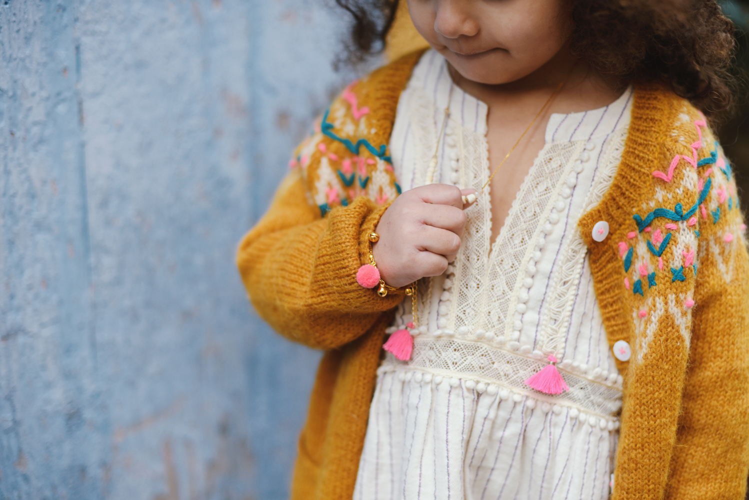 kidsbraceletkamilfluopink3 Kindermode, gelbe Strickjacke mit buntem Muster, cremefarbenes Kleid mit Spitze, rosa Quasten, Armband mit Pompon, zarte Halskette.