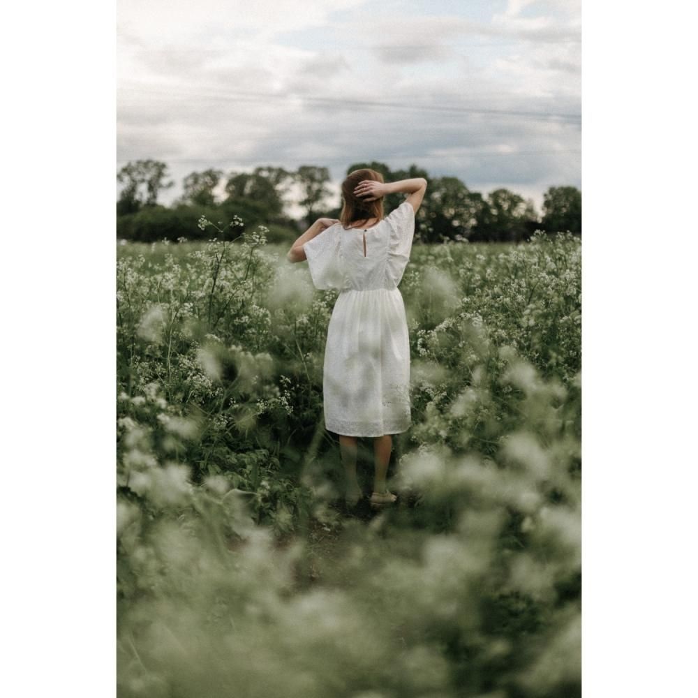 405431_large_lpxJGZMJG68ich93nGnM Frau in weißem, knielangem Kleid mit kurzen Ärmeln. Gesicht abgewandt, Hände im Haar. Steht im Feld mit weißen Blumen.