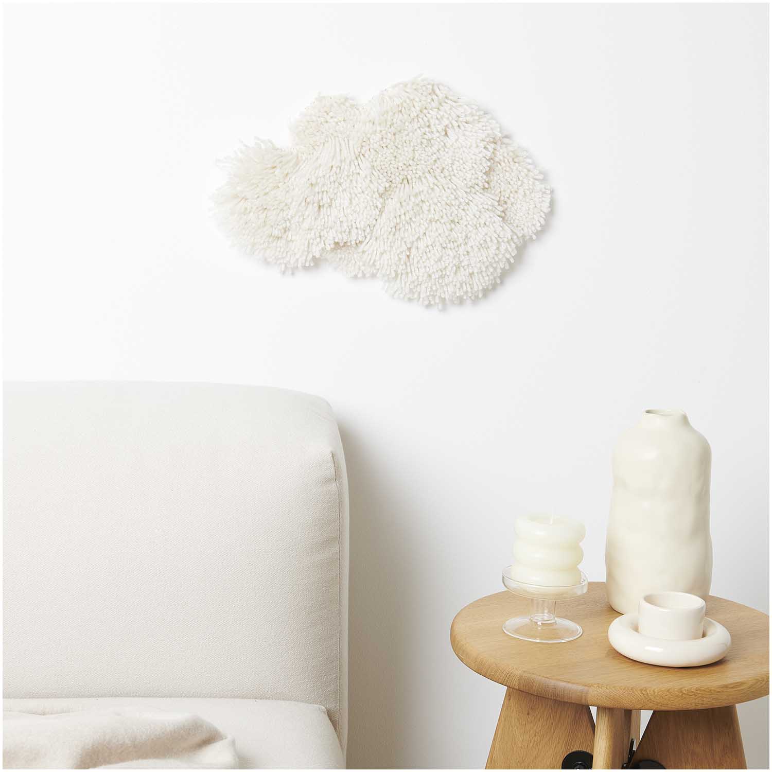 Weißes Textil-Wanddekor, unregelmäßige Form, strukturierte Oberfläche; cremefarbenes Sofa, Beistelltisch aus Holz mit cremefarbener Vase, stapelbare runde Kerzen und Tassen im minimalistischen Design.